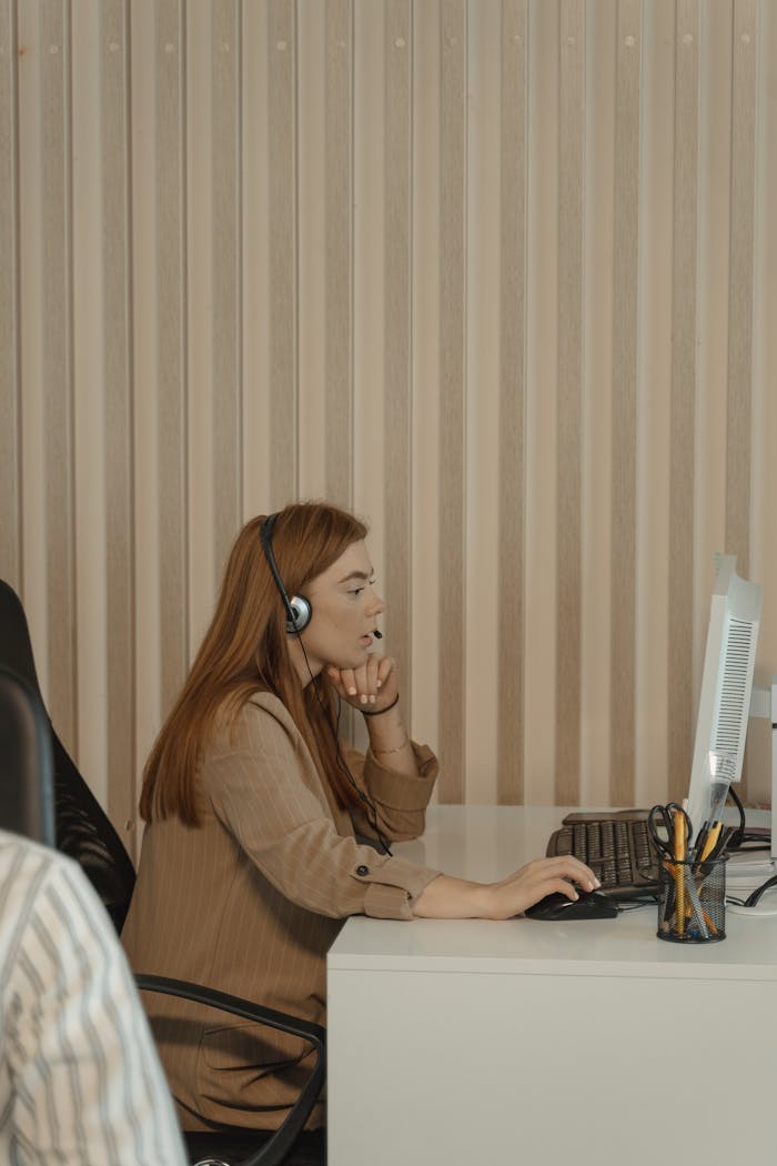 Red-haired woman with headset working at computer desk in office setting.